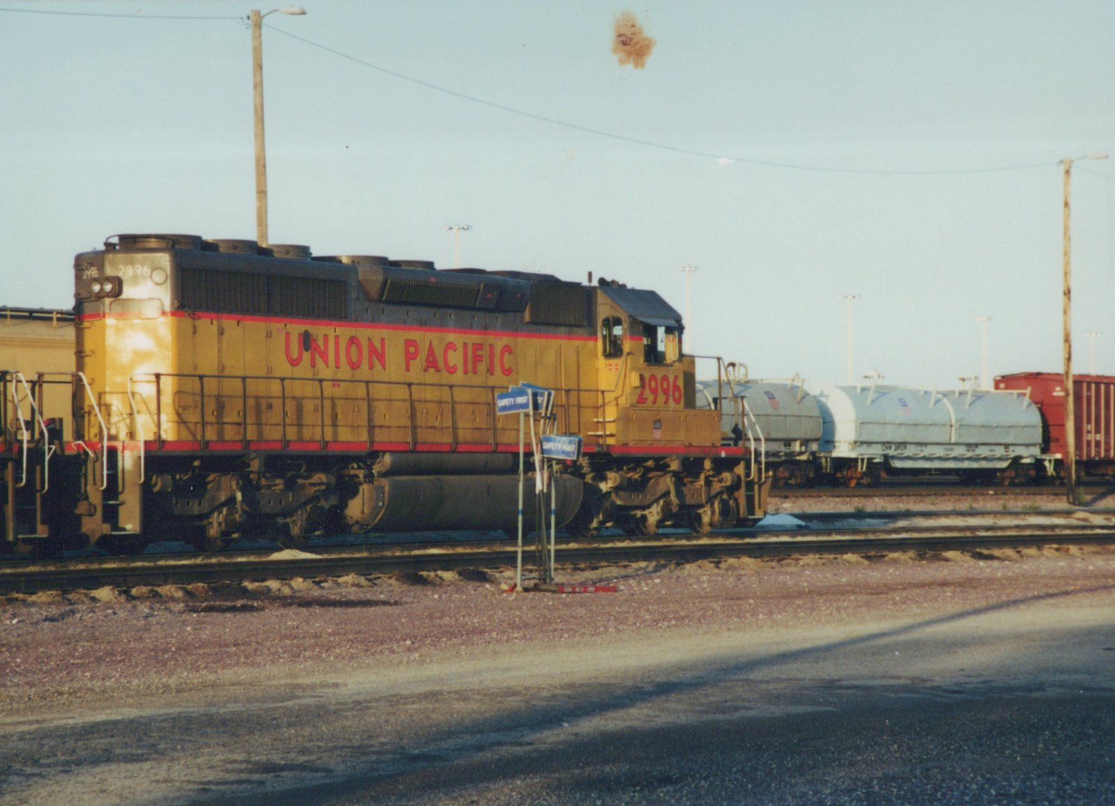 A yellow Union Pacific locomotive numbered 2996 sits on railroad tracks in a yard, with tank and boxcars visible behind it.