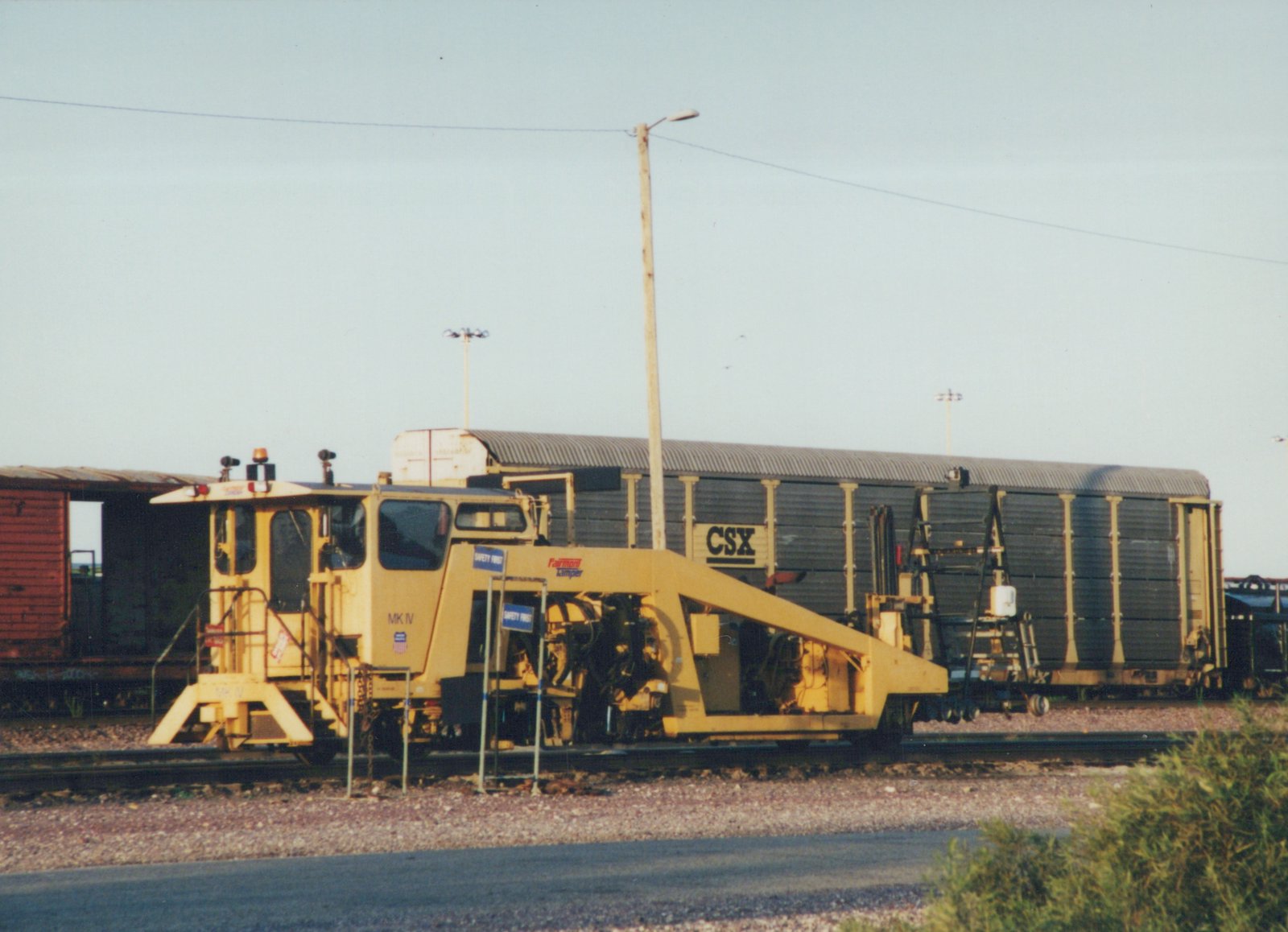 A yellow rail tamper machine marked 'MK IV' and 'Fairmont Tamper' sits on tracks beside a dark CSX boxcar under a clear sky.