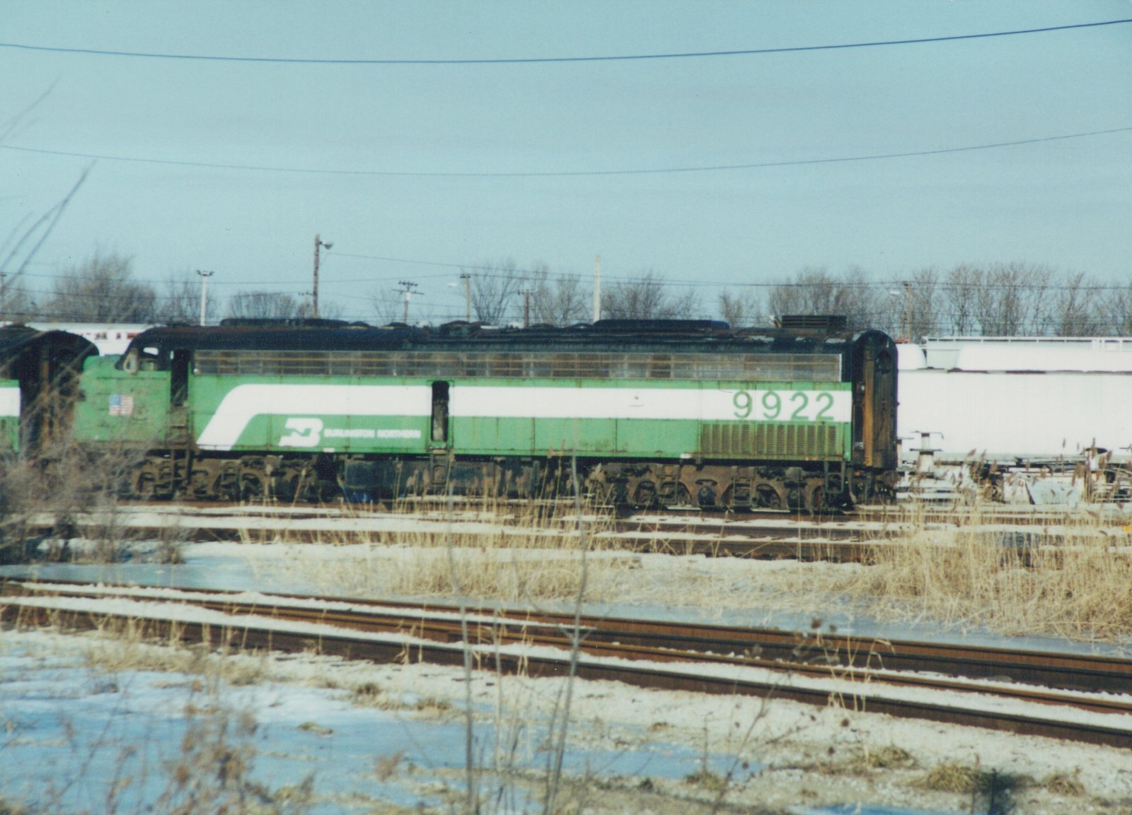 Side view of a green and white Burlington Northern locomotive numbered 9922, standing on tracks in a snowy, overcast yard with bare trees in the background.