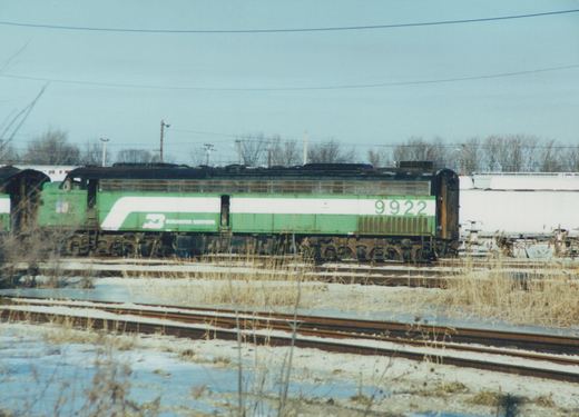 Side view of a green and white Burlington Northern locomotive numbered 9922, standing on tracks in a snowy, overcast yard with bare trees in the background.