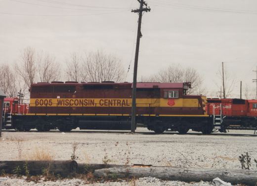 Side view of a maroon and yellow Wisconsin Central diesel locomotive with the number 6005 on its side, parked in a gravel yard with bare trees in the background.
