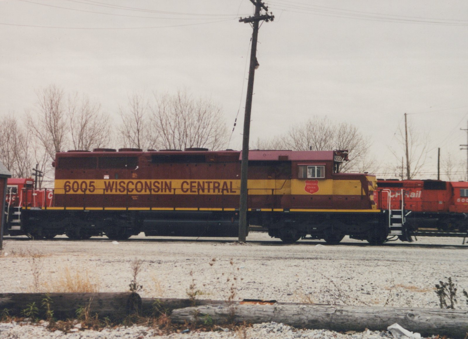 Side view of a maroon and yellow Wisconsin Central diesel locomotive with the number 6005 on its side, parked in a gravel yard with bare trees in the background.