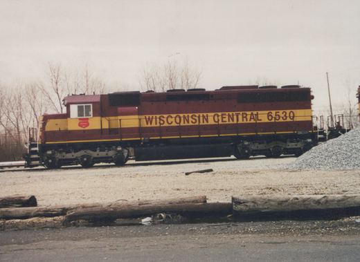 A maroon and yellow diesel locomotive with the words 'Wisconsin Central 6530' on its side, parked in a gravel rail yard with bare trees in the background, under an overcast sky.