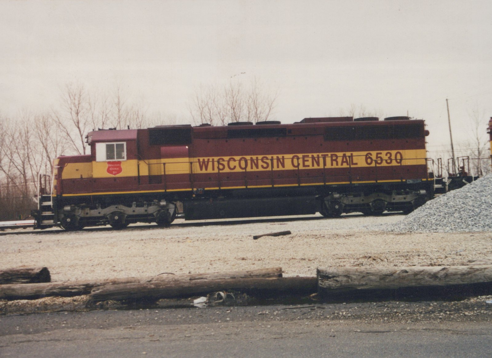 A maroon and yellow diesel locomotive with the words 'Wisconsin Central 6530' on its side, parked in a gravel rail yard with bare trees in the background, under an overcast sky.