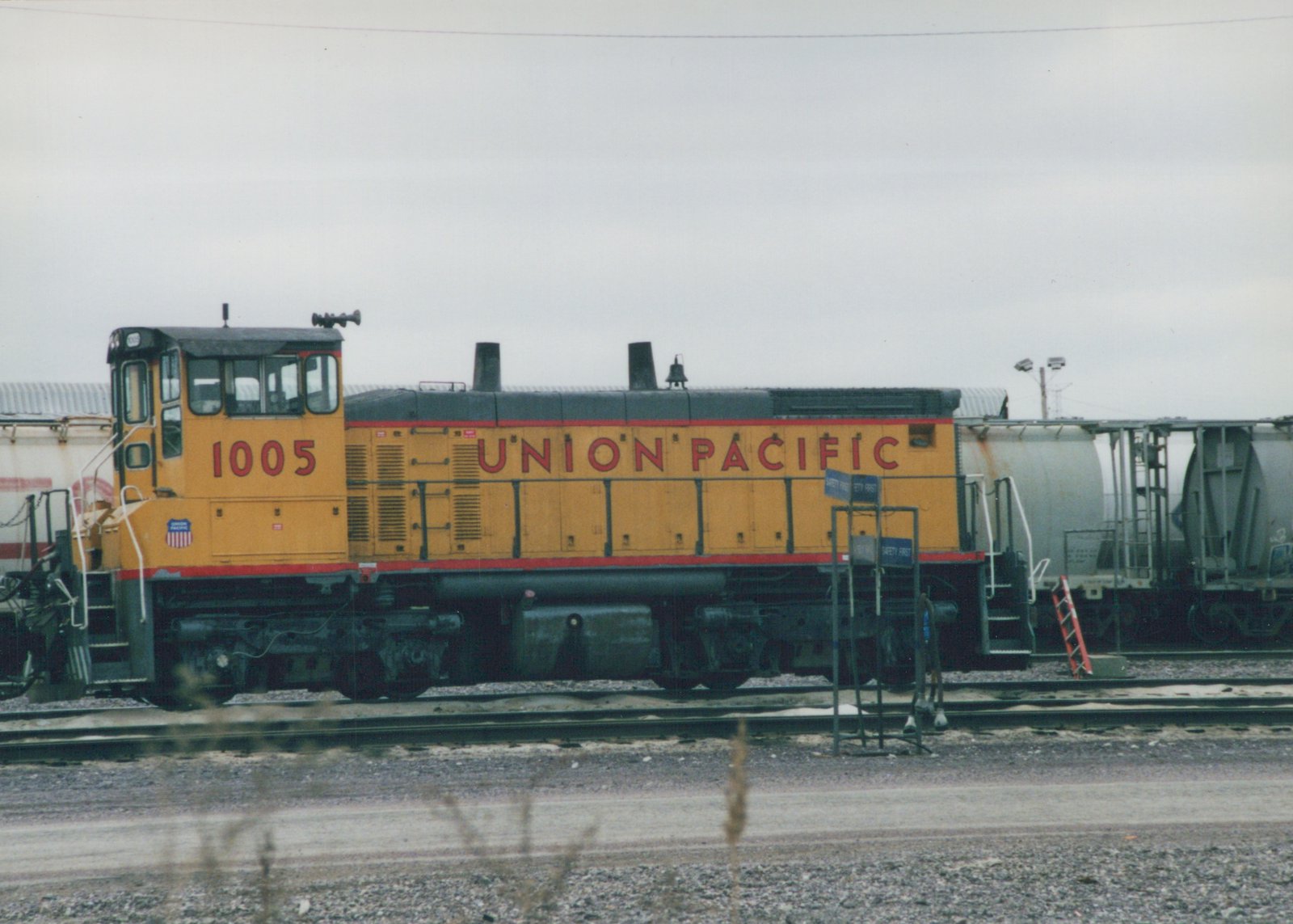 A yellow and black Union Pacific diesel locomotive numbered 1005 is parked on railroad tracks in a yard, with freight cars and a sign reading 'SAFETY FIRST' in the background.