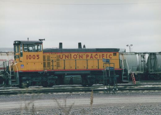 A yellow and black Union Pacific diesel locomotive numbered 1005 is parked on railroad tracks in a yard, with freight cars and a sign reading 'SAFETY FIRST' in the background.