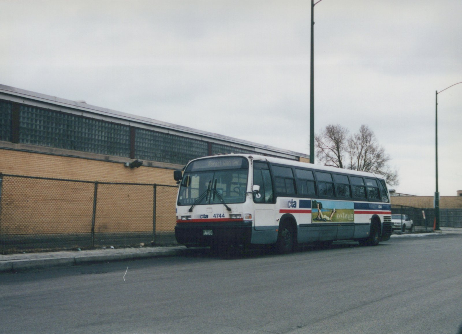 A white and blue Chicago Transit Authority bus with the number 4744 parked on a street next to a chain-link fence and a brick building with glass-block windows, under a cloudy sky.