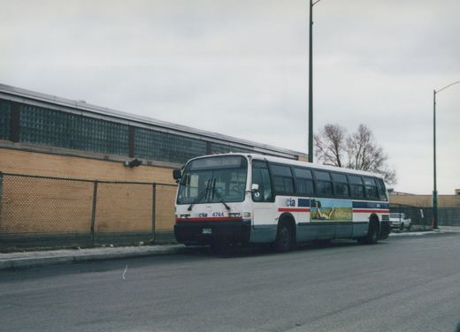A white and blue Chicago Transit Authority bus with the number 4744 parked on a street next to a chain-link fence and a brick building with glass-block windows, under a cloudy sky.