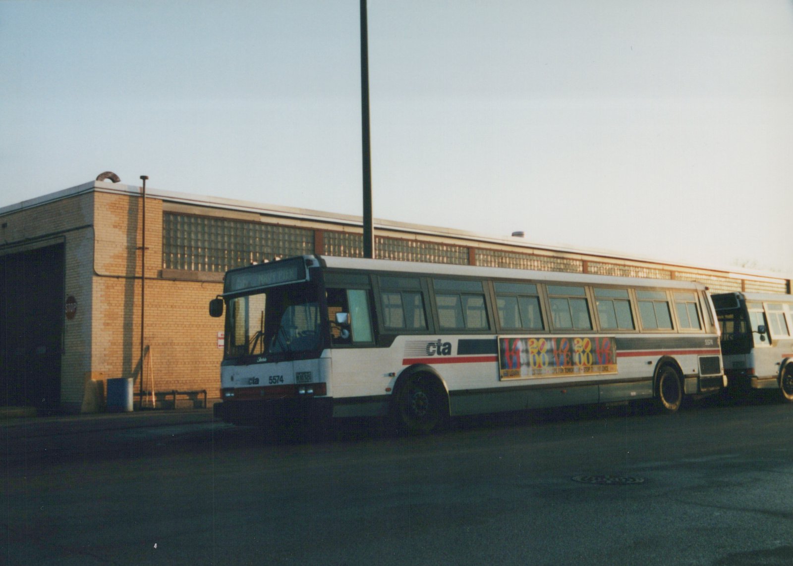 A white CTA bus with red and blue stripes, number 5574, parked on a street in front of a brick building with glass block windows, facing slightly left with 'NAVY PIER' on its destination sign.