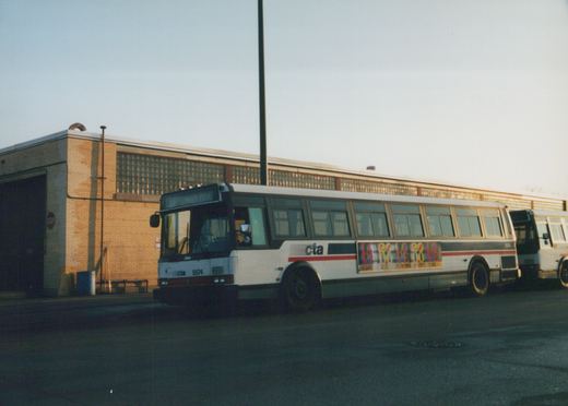 A white CTA bus with red and blue stripes, number 5574, parked on a street in front of a brick building with glass block windows, facing slightly left with 'NAVY PIER' on its destination sign.