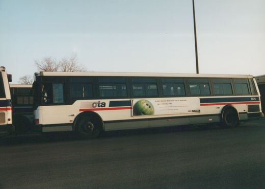 Side view of a white CTA bus with blue and red stripes, number 5575, showing an anti-drug ad with a bowling ball, parked in a lot with bare trees in the background.