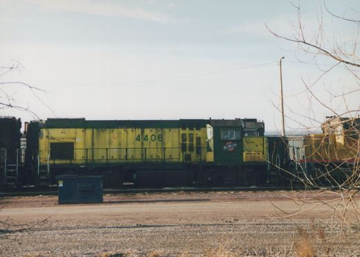 A side view of a Chicago and North Western yellow and green diesel locomotive number 4406, parked in a gravel yard with bare trees in the foreground and another locomotive partially visible in the background.