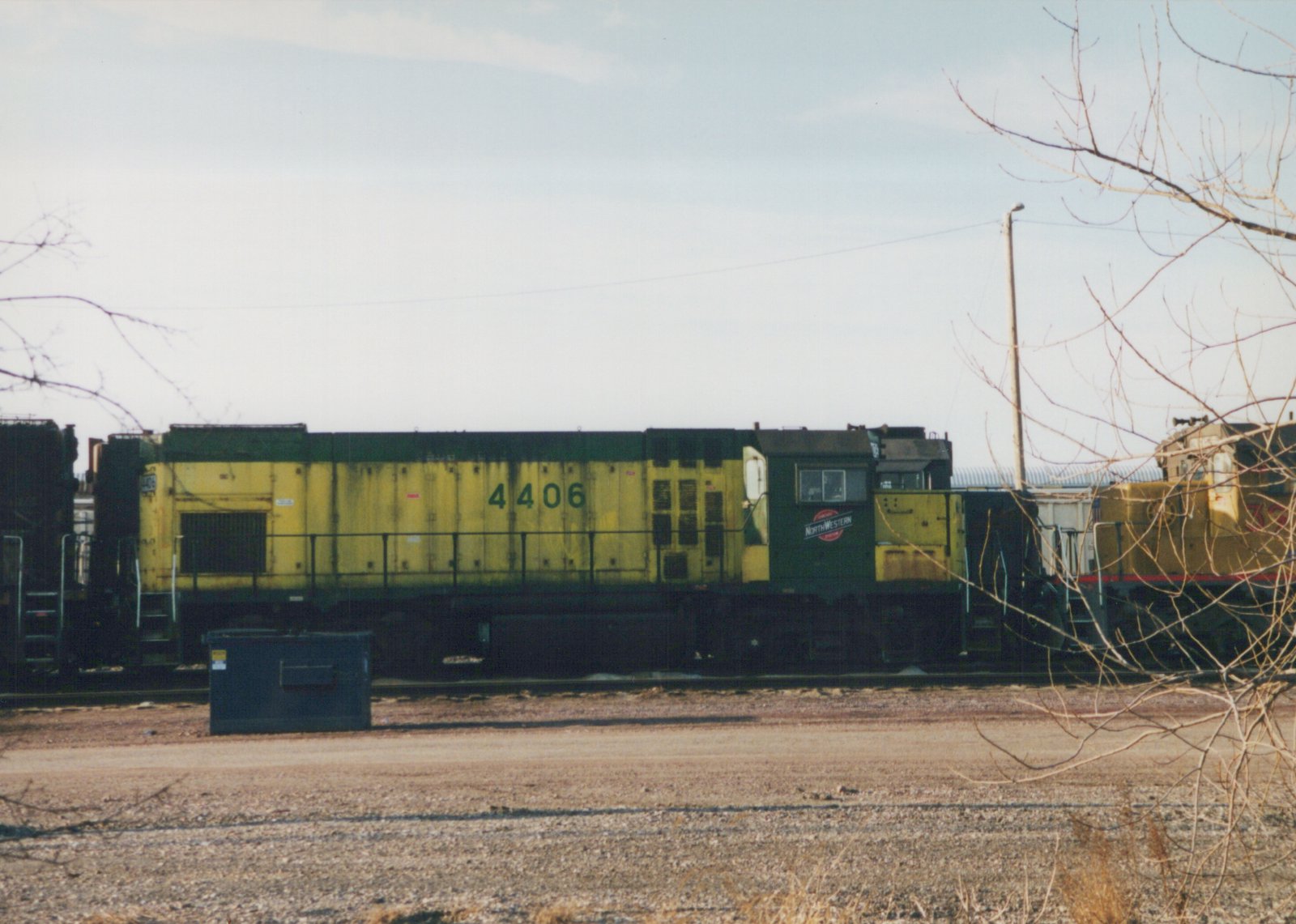A side view of a Chicago and North Western yellow and green diesel locomotive number 4406, parked in a gravel yard with bare trees in the foreground and another locomotive partially visible in the background.