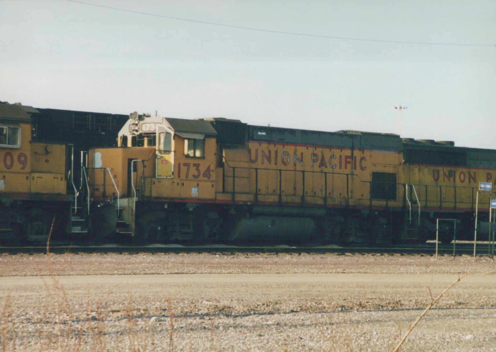 Side view of a yellow Union Pacific diesel locomotive, number 1734, standing on tracks in a gravel yard with a second locomotive partially visible behind it.