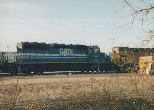A blue GATX locomotive with the number 7361 on its side sits in a rail yard, with a yellow Union Pacific locomotive in the background and bare branches framing the scene.