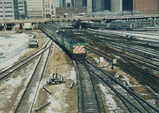 A green Metra diesel locomotive numbered 9019 moves through a snow-dusted rail yard with multiple tracks and Amtrak passenger cars in the background.