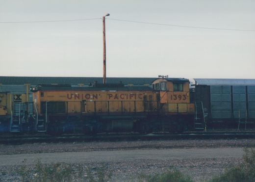 A side view of a yellow Union Pacific locomotive numbered 1393, with a freight car behind it, on a mainline track in a rail yard under an overcast sky.