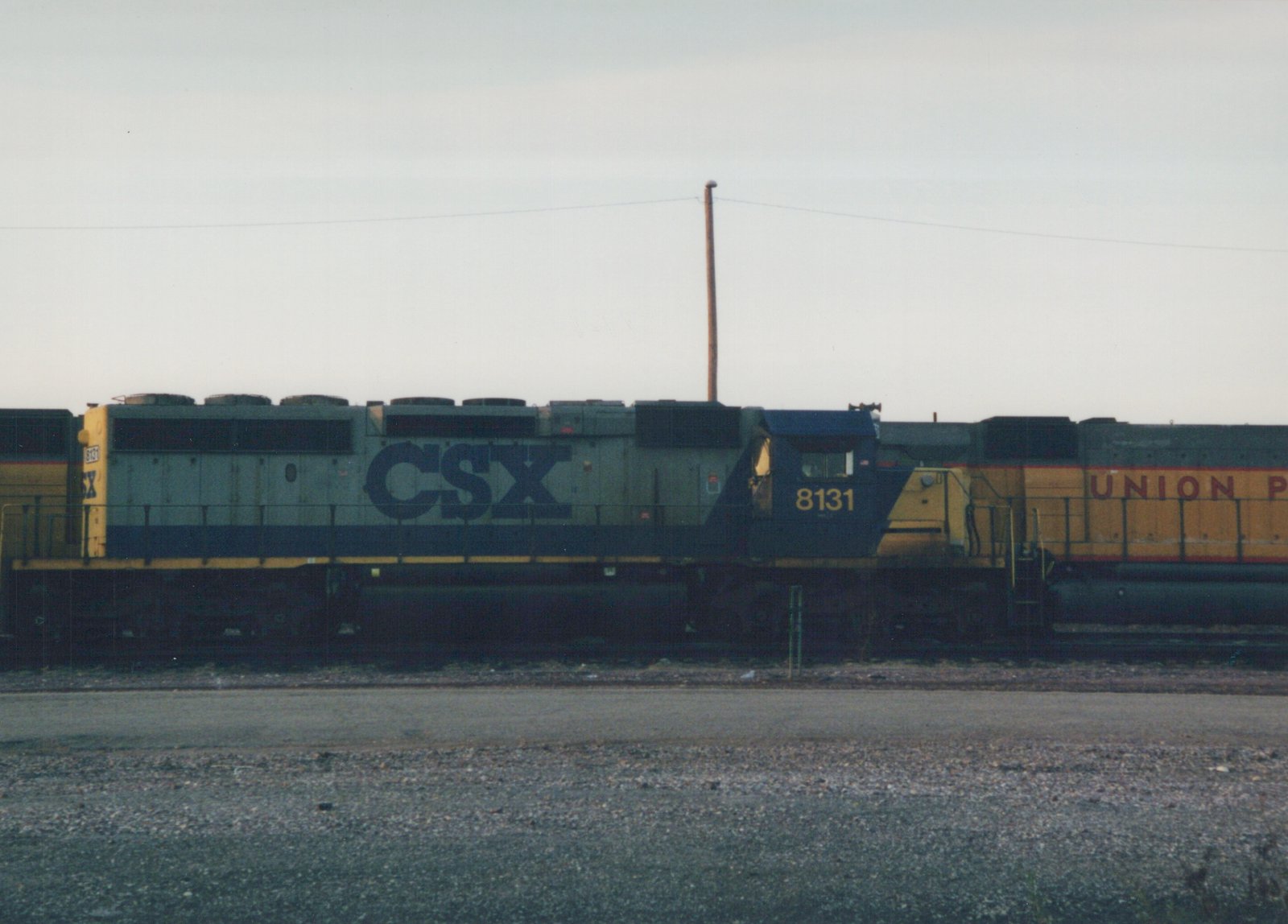 A CSX diesel locomotive with the number 8131 on its side and front, parked in a rail yard with another Union Pacific locomotive partially visible behind it under an overcast sky.