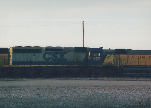 A CSX diesel locomotive with the number 8131 on its side and front, parked in a rail yard with another Union Pacific locomotive partially visible behind it under an overcast sky.