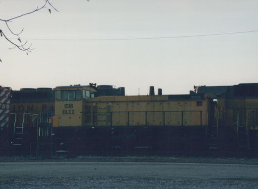 A yellow and black HLCX 1518 diesel locomotive stands in a rail yard with other locomotives under a cloudy sky.