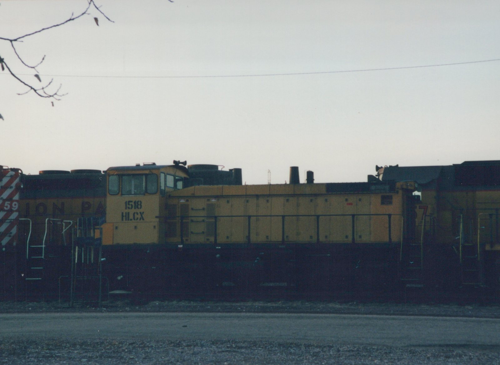 A yellow and black HLCX 1518 diesel locomotive stands in a rail yard with other locomotives under a cloudy sky.