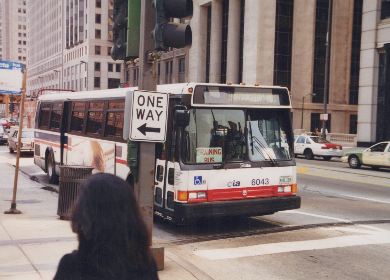 A white and red Chicago Transit Authority bus, number 6043, labeled 'INSTRUCTION BUS' and 'TRAINING BUS,' is on a city street with pedestrians and taxis in the background.