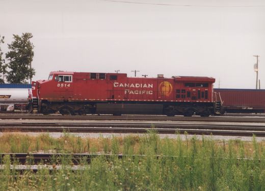 A red Canadian Pacific diesel locomotive numbered 8514 is shown on a railroad track with freight cars, under an overcast sky, with grass in the foreground.