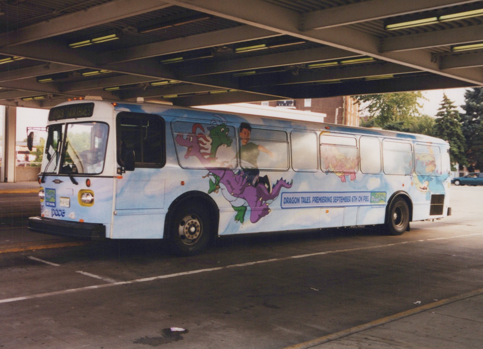 A light blue Pace bus with a colorful 'Dragon Tales' cartoon ad on its side is parked under a concrete overpass, showing its front and side.