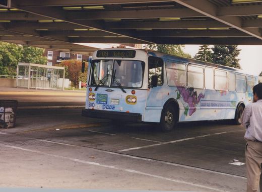 A light blue Pace bus with a 'Dragon Tales' cartoon wrap is parked under a concrete overpass. The front display shows route 209 to the Transit Center. A person stands nearby.