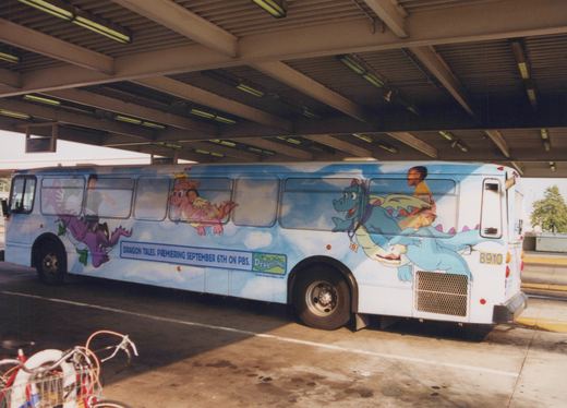 A side view of a white city bus parked under a shelter, covered in a 'Dragon Tales' cartoon advertisement with dragons and children, showing fleet number 8910 on the rear.