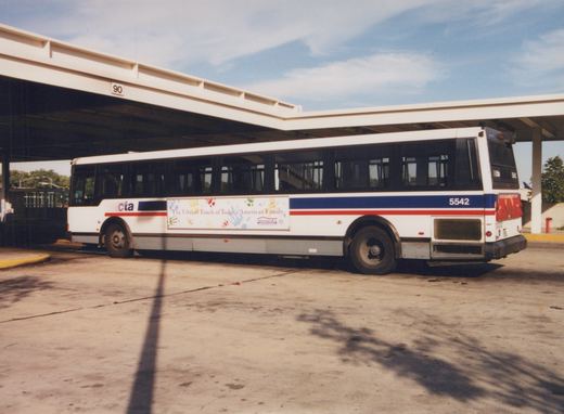 A white, blue, and red Chicago Transit Authority bus, number 5542, is parked at a bus terminal under a concrete shelter on a clear day.