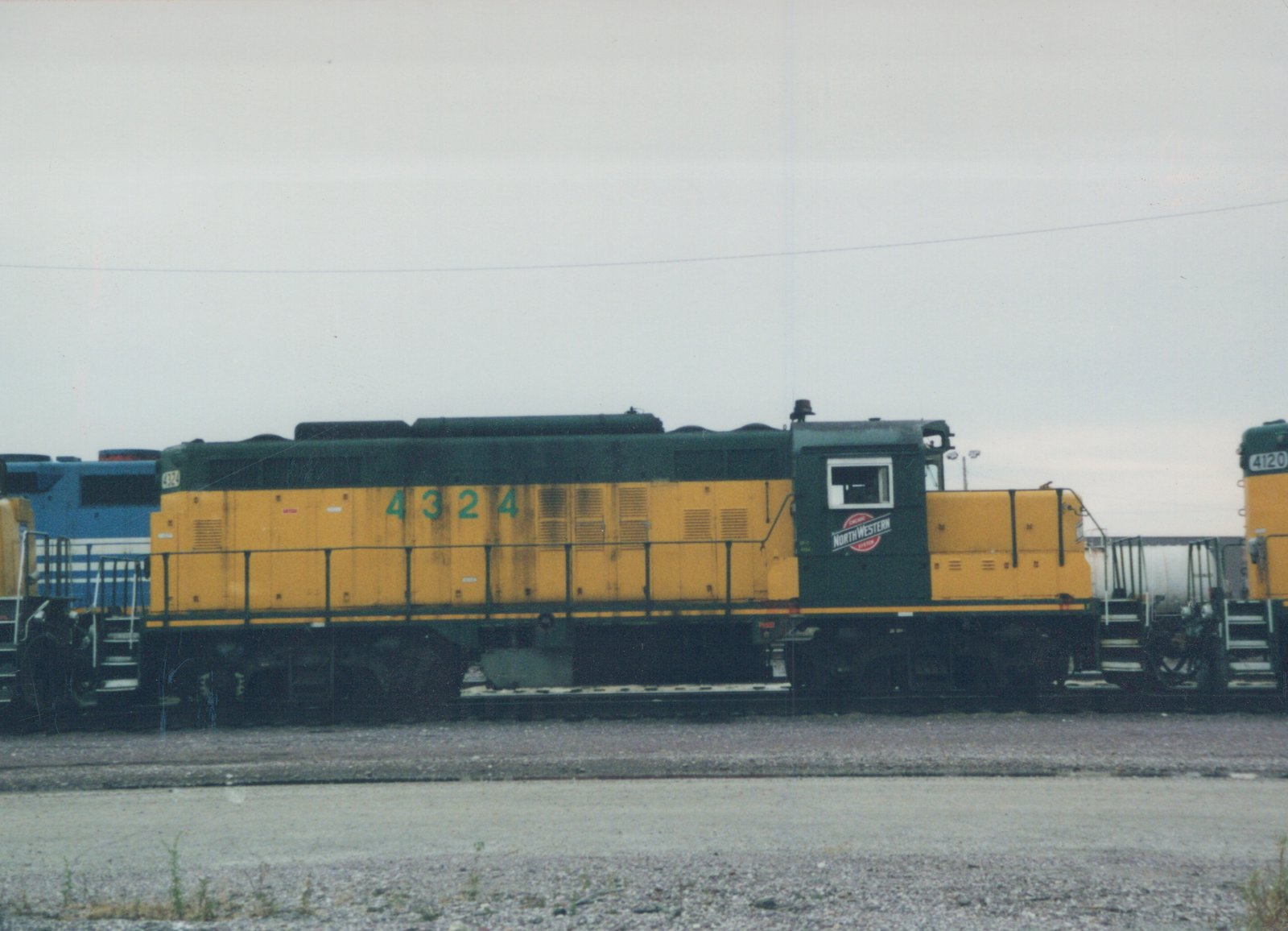 Side view of a green and yellow CNW diesel locomotive numbered 4324 parked on tracks in a yard with other locomotives nearby under an overcast sky.