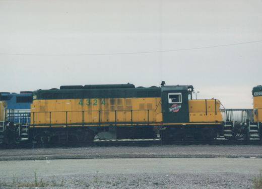 Side view of a green and yellow CNW diesel locomotive numbered 4324 parked on tracks in a yard with other locomotives nearby under an overcast sky.