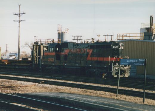 A black and red diesel locomotive with the number 2923 and 'HARBOR' lettering is parked at a station next to a sign reading 'Mannhiem Platform 1' with directions to and from Chicago.