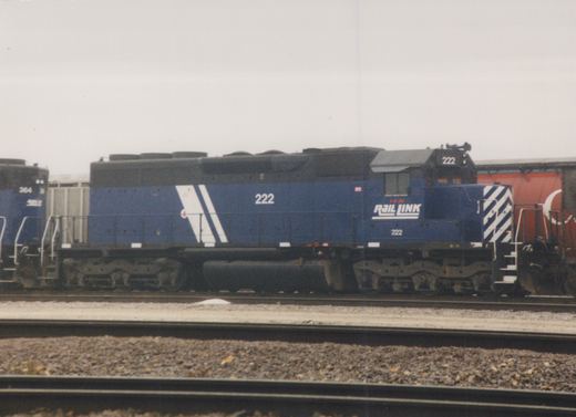 A side view of a blue Rail Link diesel locomotive numbered 222, parked on railroad tracks with another similar locomotive, number 364, visible in the background under an overcast sky.