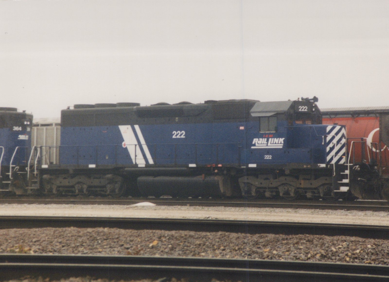 A side view of a blue Rail Link diesel locomotive numbered 222, parked on railroad tracks with another similar locomotive, number 364, visible in the background under an overcast sky.