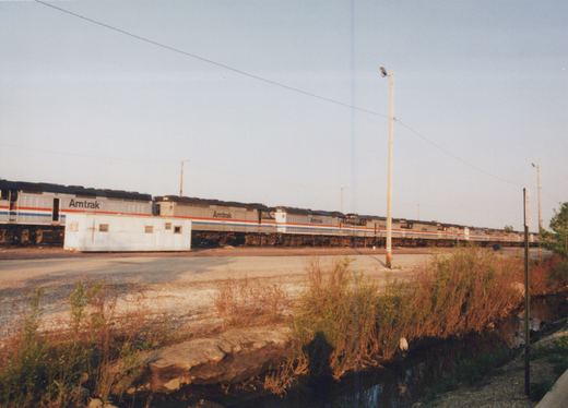 A line of silver Amtrak locomotives with red and blue stripes sits at a rail yard, with a small white building in the foreground and brush and a drainage ditch in the foreground.