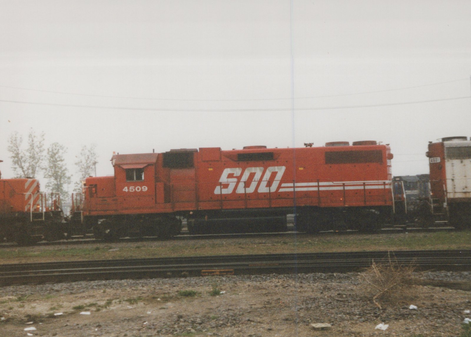 A side view of a bright orange Soo Line locomotive numbered 4509 parked on a set of tracks in a yard on an overcast day, with another locomotive visible behind it.