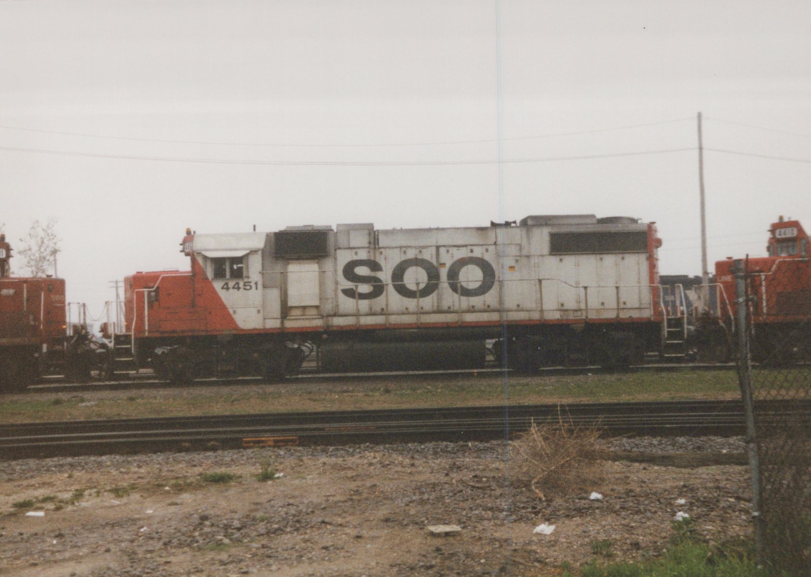 A side view of a white and orange Soo Line diesel locomotive, number 4451, sitting on tracks in a rail yard with a second locomotive visible behind it, under a gray overcast sky.