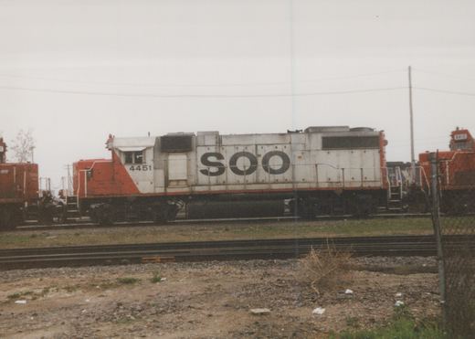 A side view of a white and orange Soo Line diesel locomotive, number 4451, sitting on tracks in a rail yard with a second locomotive visible behind it, under a gray overcast sky.