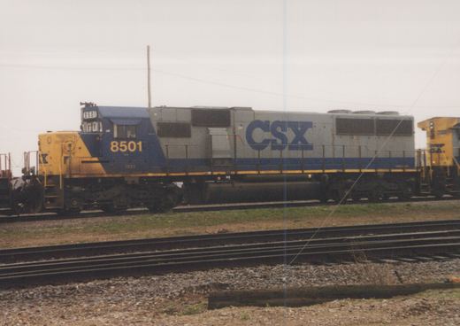 A side view of a CSX diesel locomotive numbered 8501, painted in blue, gray, and yellow, sitting on tracks in a gravel yard under an overcast sky.