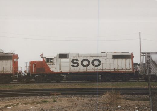 A side view of a white and orange Soo Line diesel locomotive, number 4410, parked in a rail yard next to a freight car.