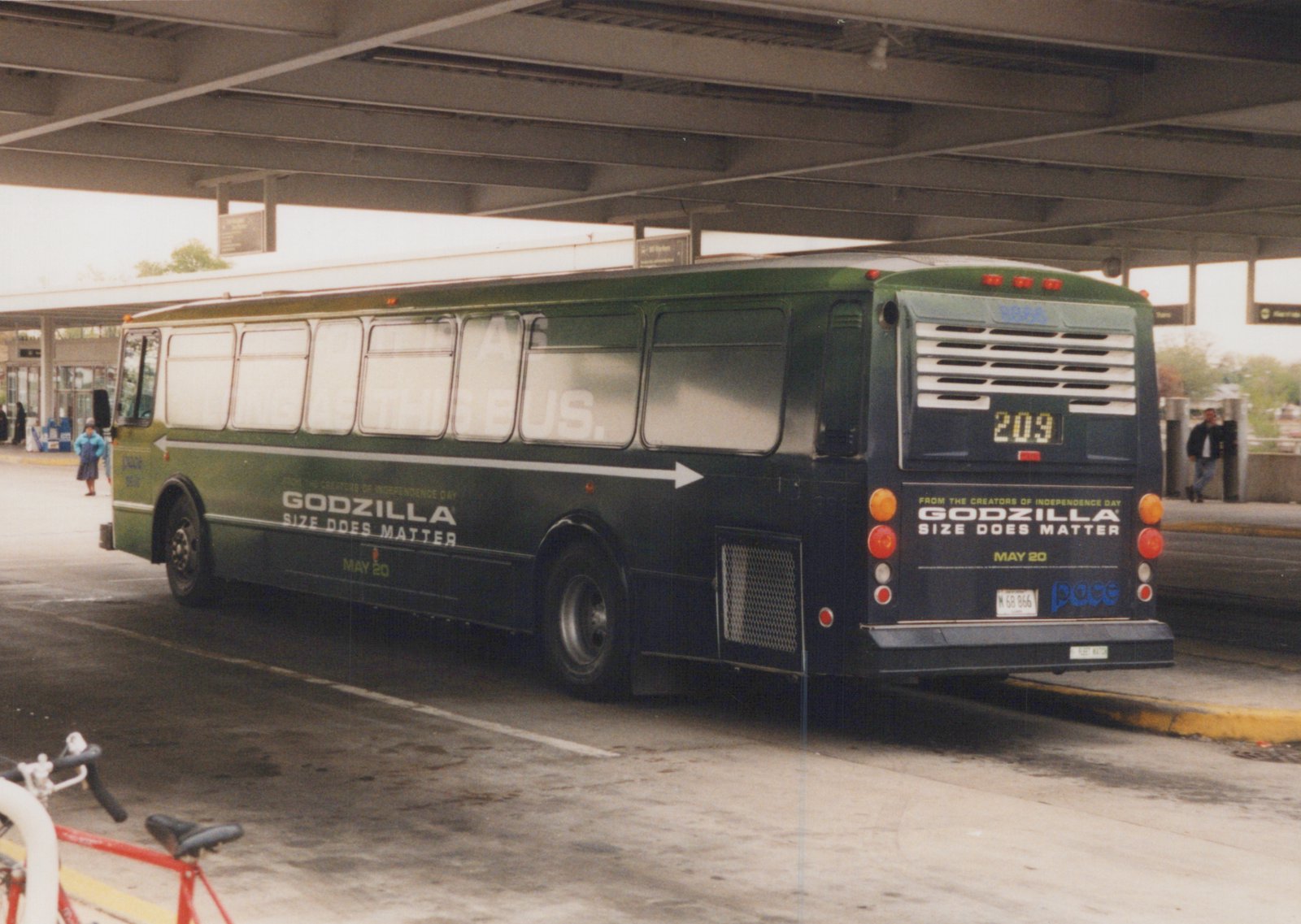 A green Pace motor bus with a Godzilla movie ad parked under an elevated highway, showing its rear and side.