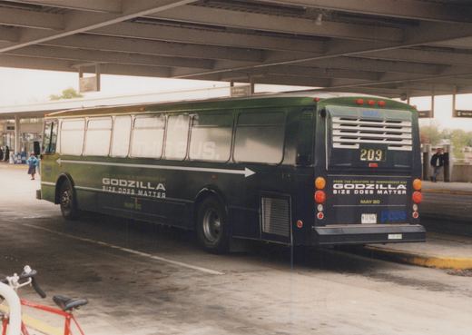 A green Pace motor bus with a Godzilla movie ad parked under an elevated highway, showing its rear and side.