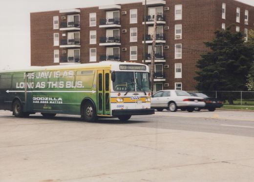 A white and yellow Pace bus with a green Godzilla advertisement on its side, number 8866, on a street in front of a brick apartment building.