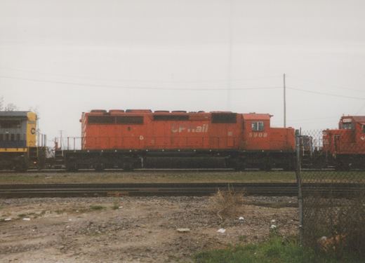 Side view of a bright orange CP Rail diesel locomotive, number 5908, parked on a track in a gravel yard with a chain-link fence and another locomotive visible in the background.