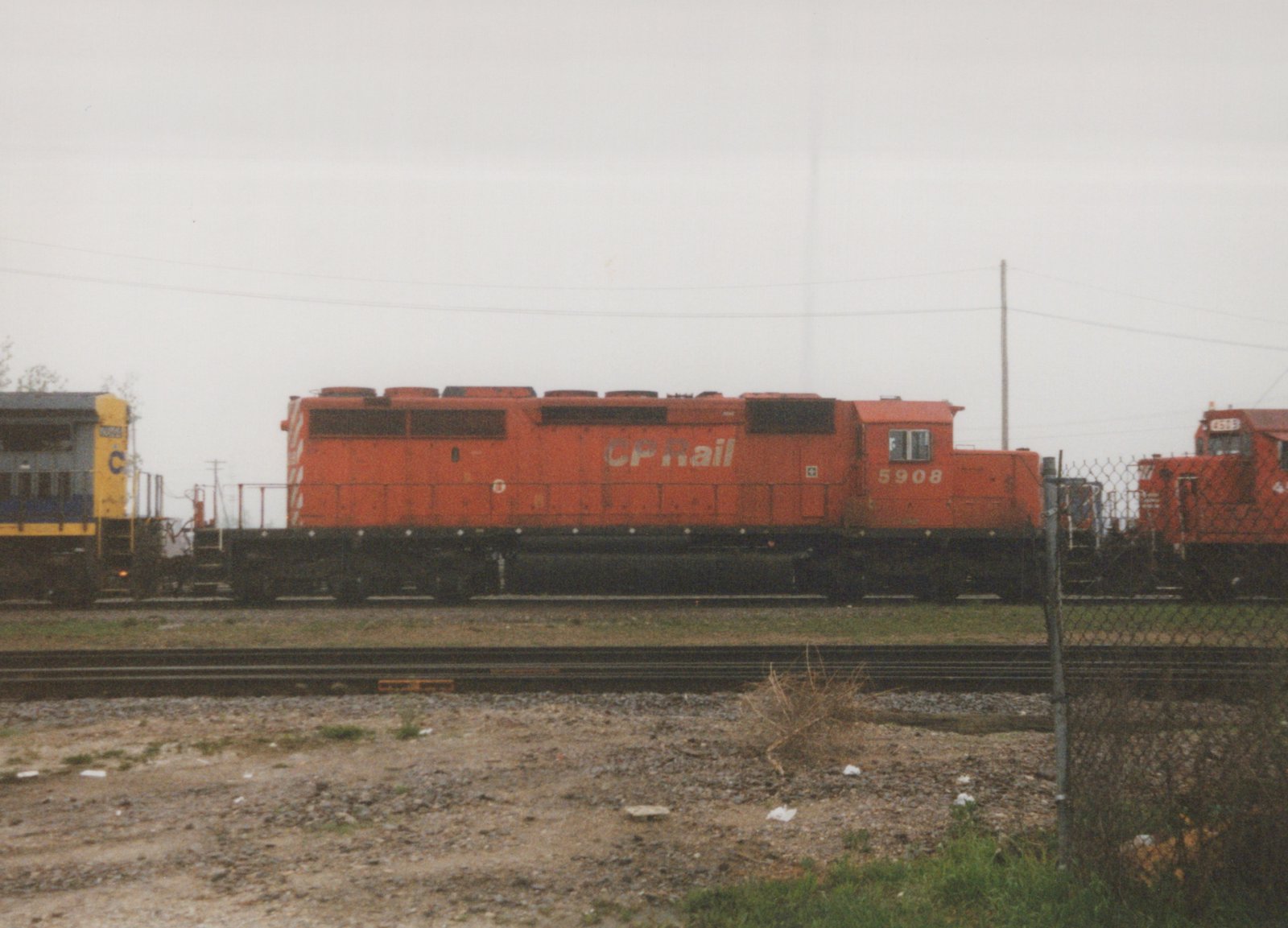 Side view of a bright orange CP Rail diesel locomotive, number 5908, parked on a track in a gravel yard with a chain-link fence and another locomotive visible in the background.