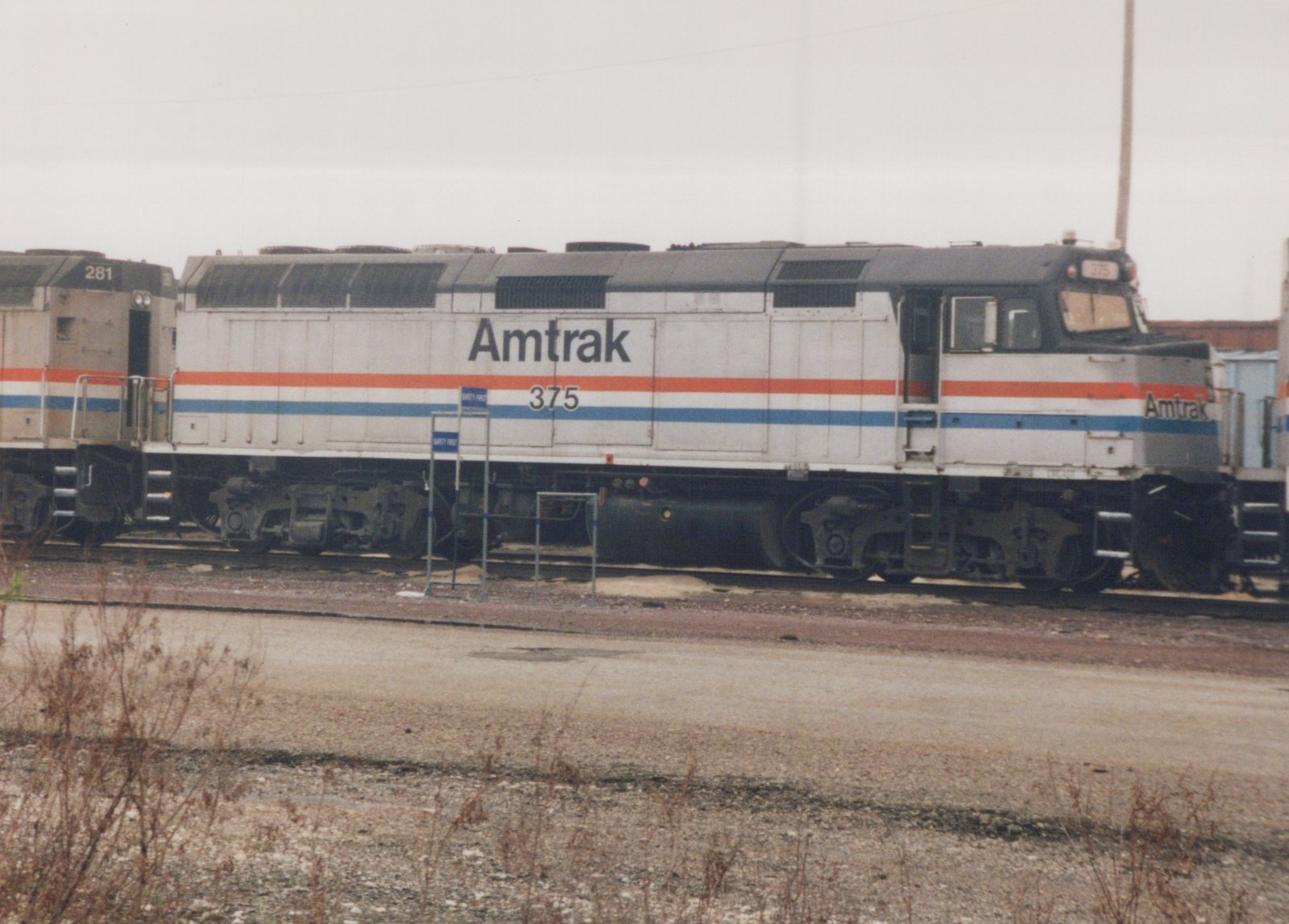 A side view of an Amtrak locomotive, number 375, in white livery with red and blue stripes, parked on tracks in a gravel yard with a second locomotive partially visible.