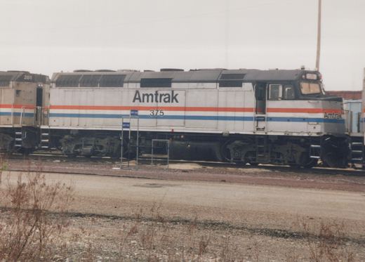 A side view of an Amtrak locomotive, number 375, in white livery with red and blue stripes, parked on tracks in a gravel yard with a second locomotive partially visible.