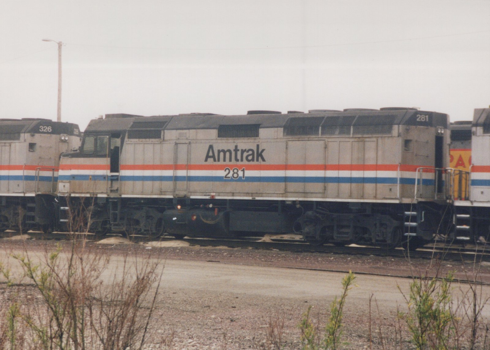A side view of an Amtrak F40PH locomotive, number 281, parked on a track in a rail yard with another Amtrak locomotive, number 326, visible behind it. The sky is overcast, and there are weeds in the foreground.
